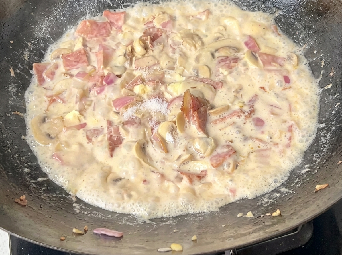Cooked spaghetti being added to a pan filled with creamy bacon and mushroom sauce.