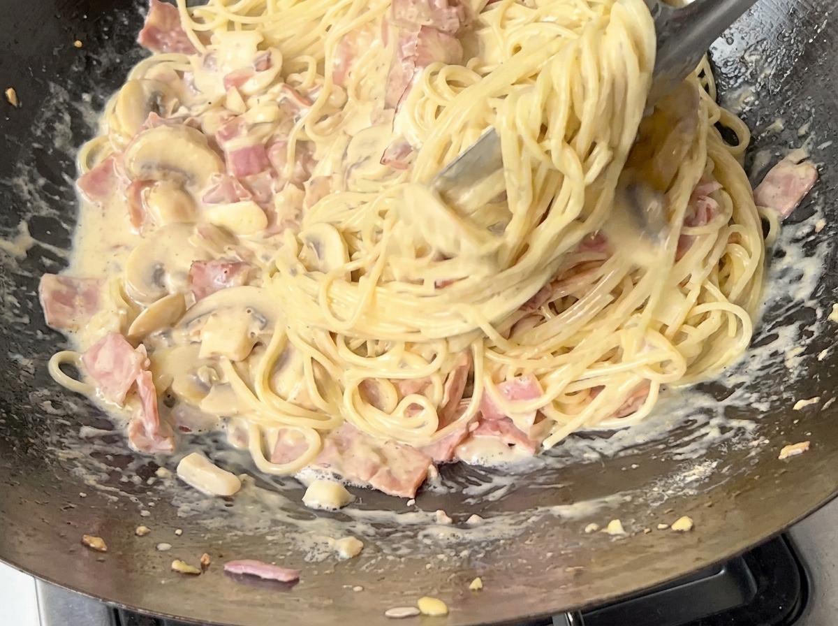 A close-up of spaghetti being tossed in a creamy bacon and mushroom sauce using metal tongs.