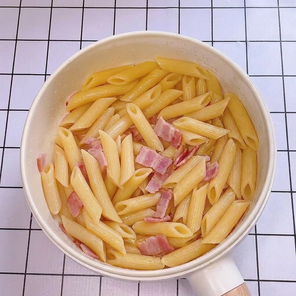 Pasta water being poured into the pan with penne and bacon, creating a glossy sheen.
