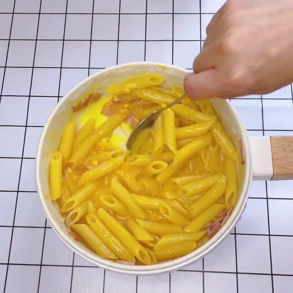 Close-up of a spoon rapidly tossing pasta and bacon in a pan with a thick, yellow, creamy egg-cheese sauce.