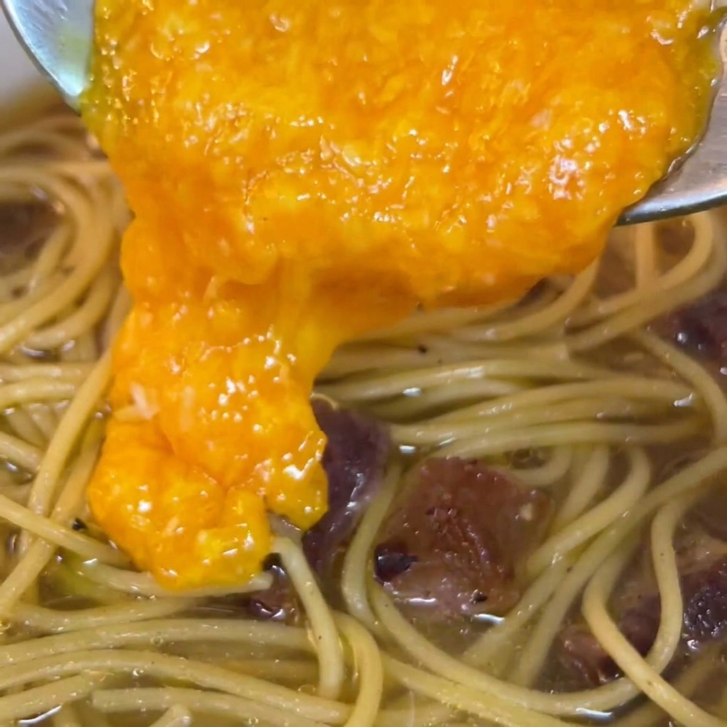 A rich, vibrant orange egg and cheese mixture being poured from a spoon onto the pasta in the pan.