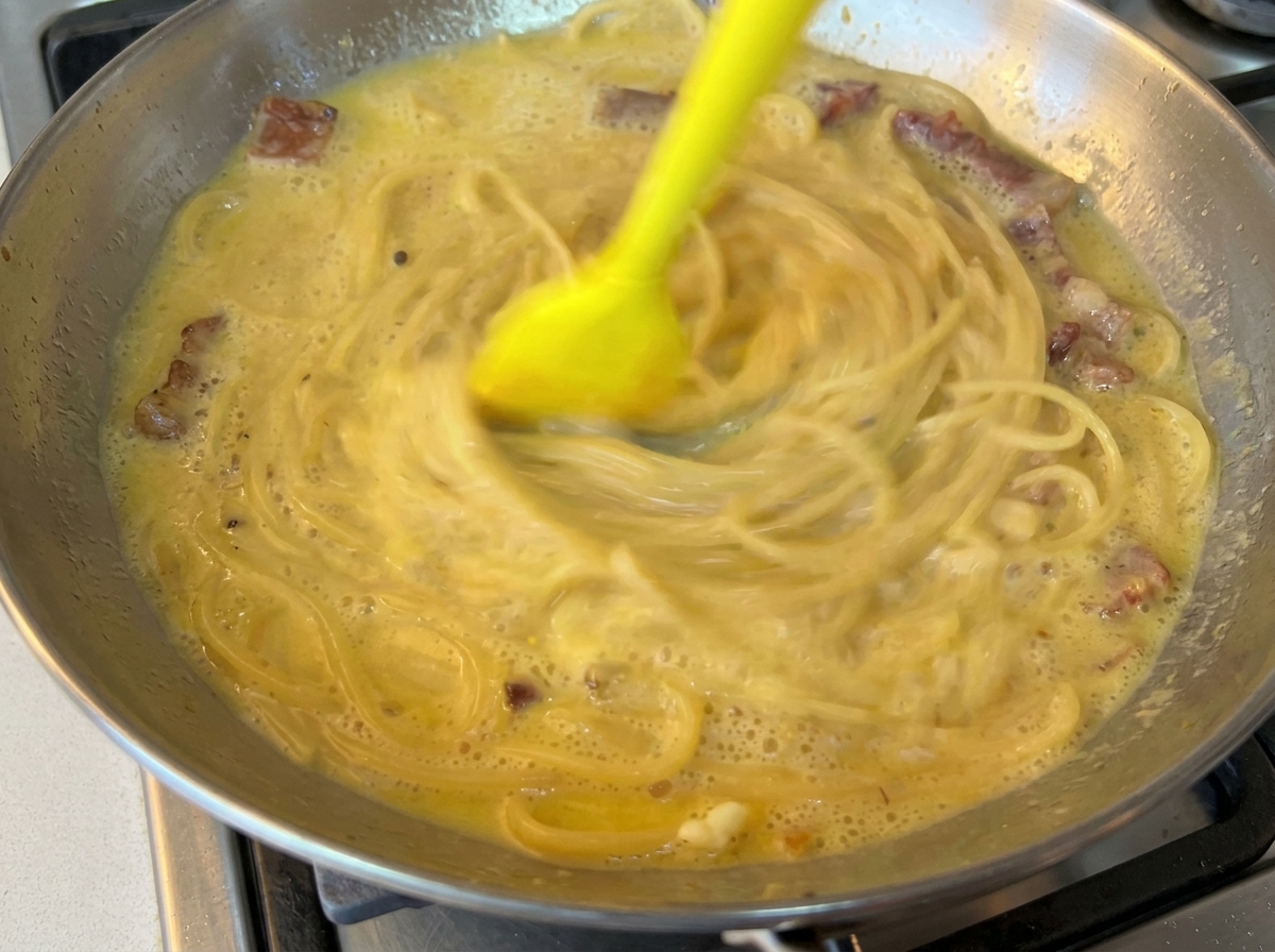 A yellow rubber spatula rapidly mixing spaghetti with a creamy egg-based sauce in a stainless steel pan.