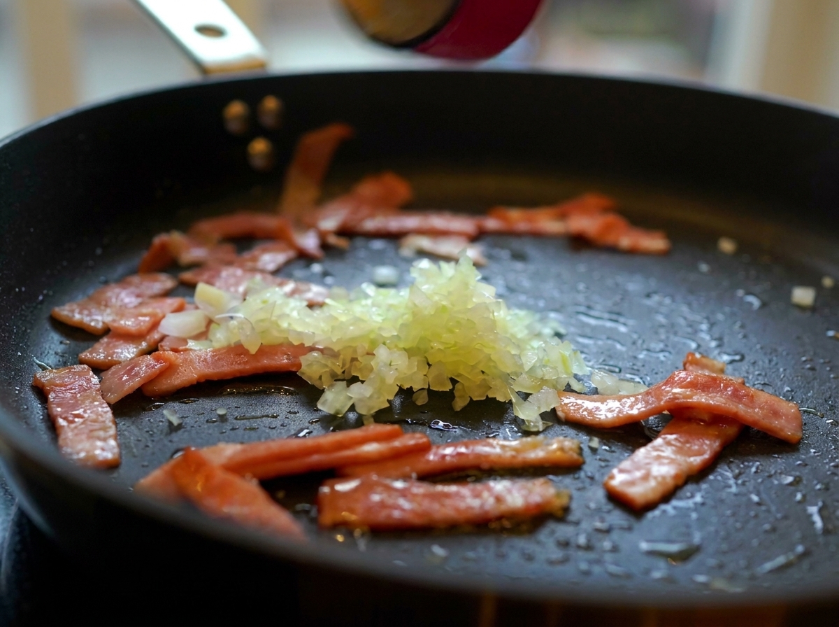 Minced onions, garlic, and sliced mushrooms being added to a pan with fried bacon.