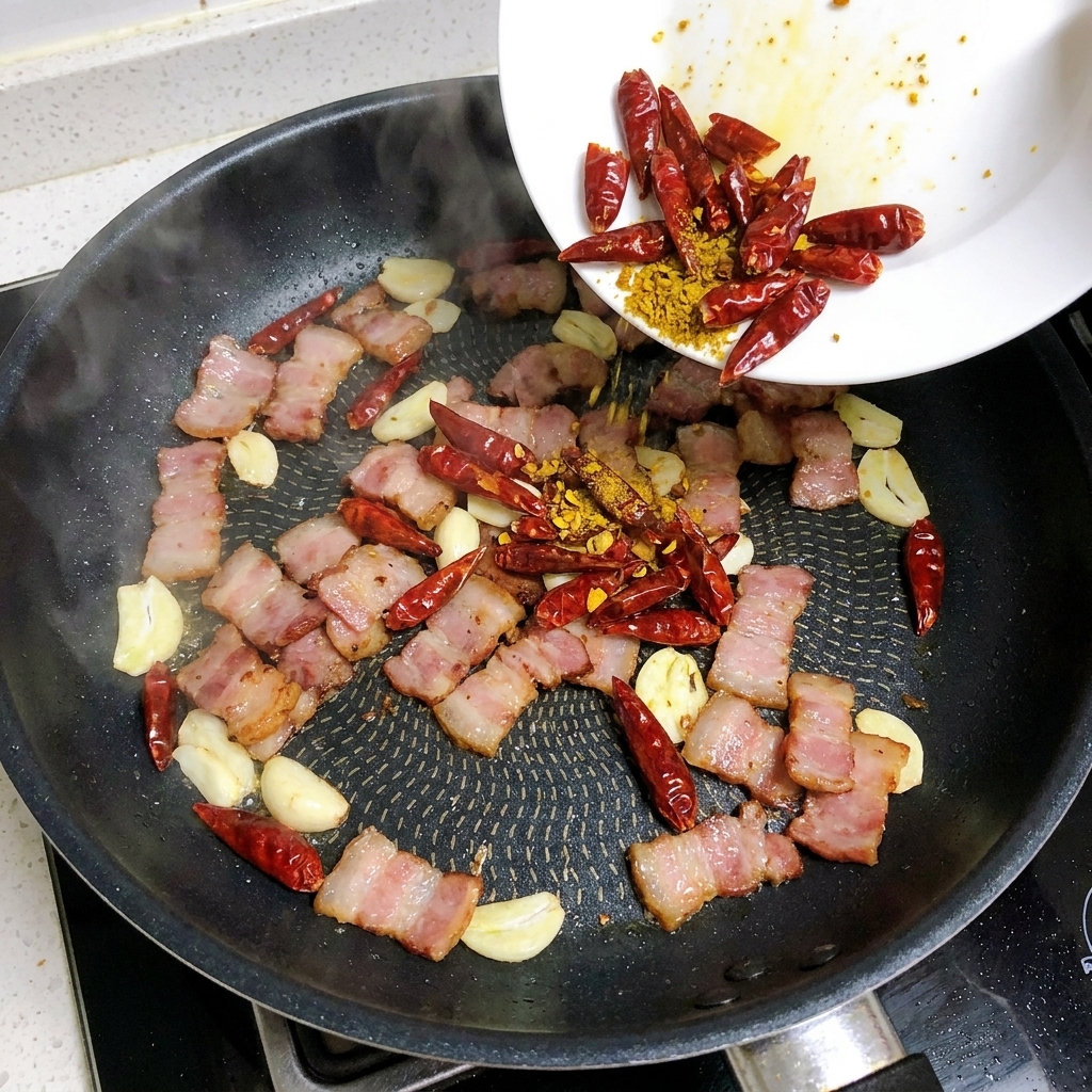 Slices of garlic and dried red chilies being added to a pan containing strips of frying bacon.