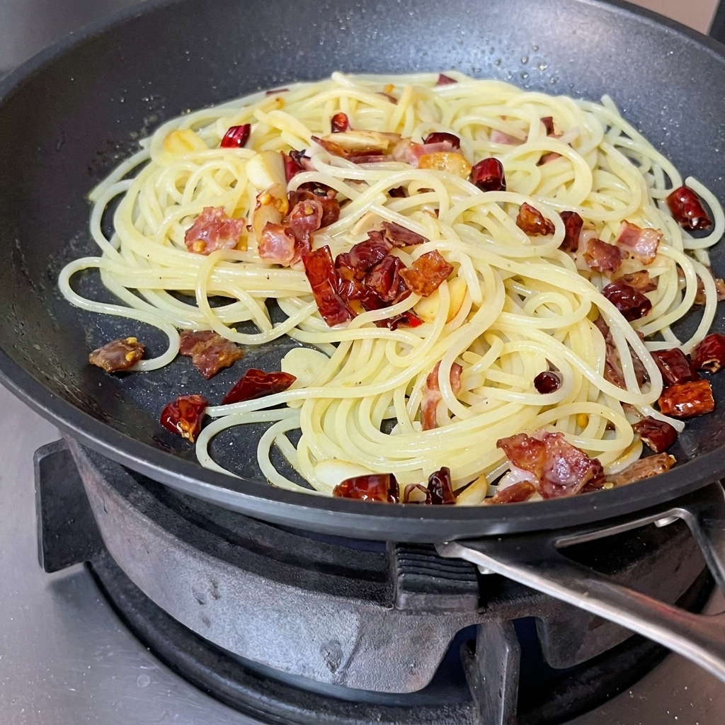 Spaghetti, bacon, and aromatics being tossed and mixed together in a pan with a red spatula.