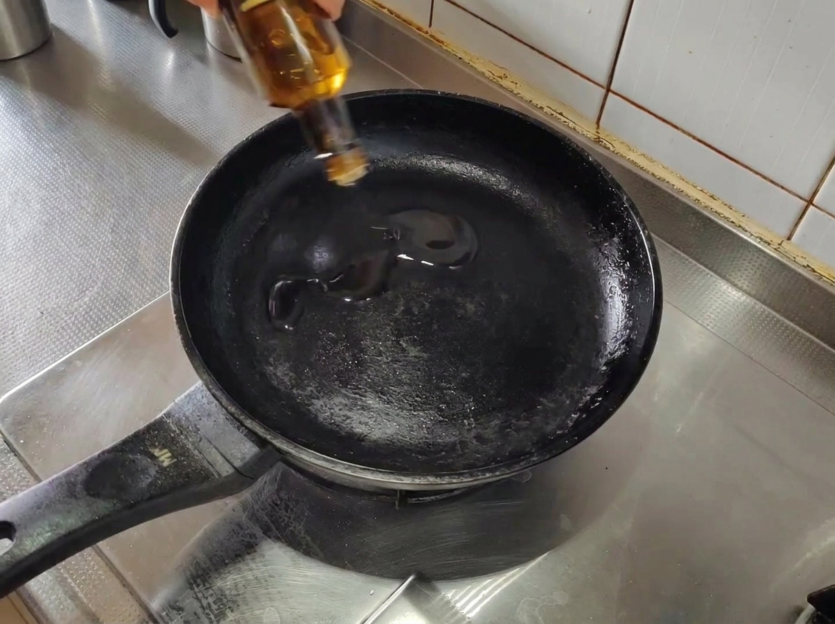Olive oil being poured into an empty, preheated black skillet on a stovetop.