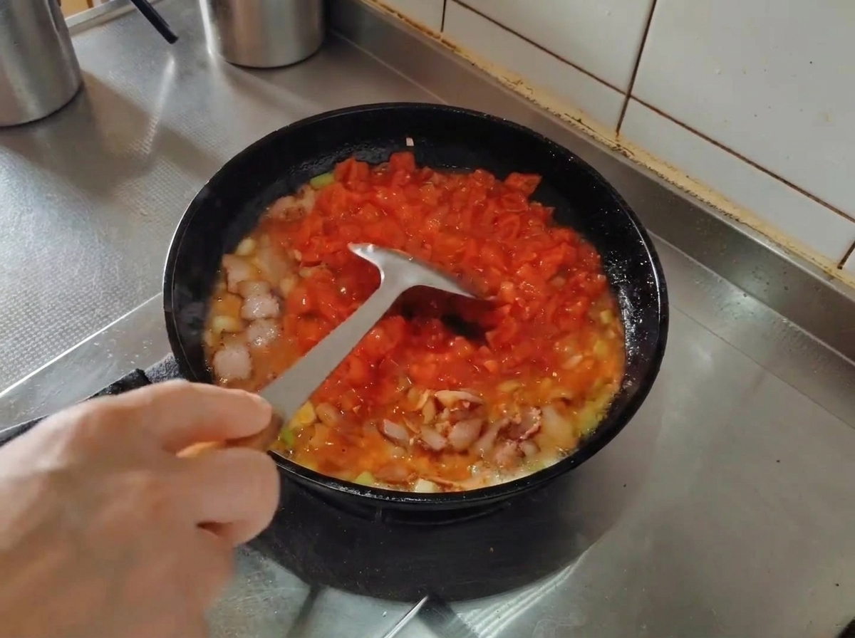 A metal spatula mashing diced tomatoes in a skillet while cooking with bacon and aromatics.