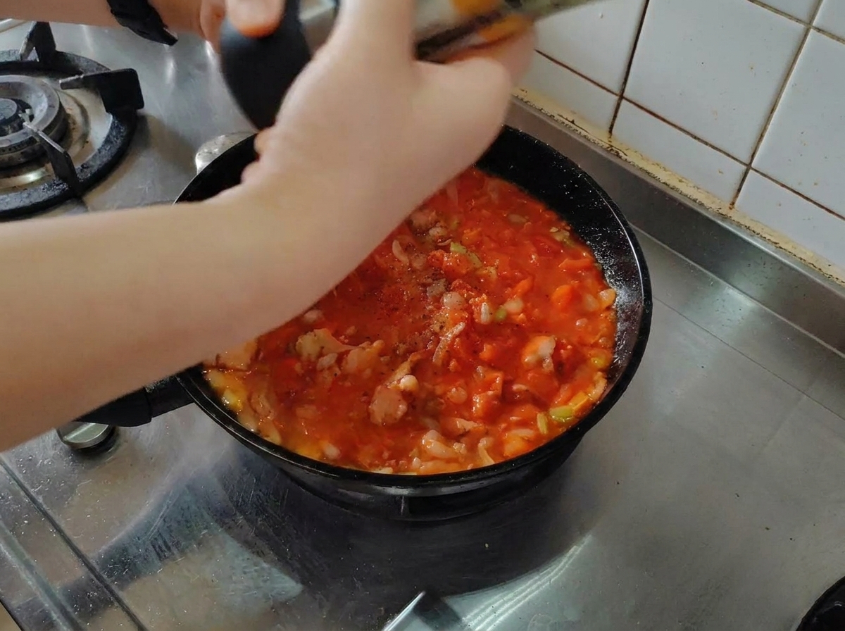Fresh black pepper being ground over a simmering tomato and bacon sauce in a black skillet.