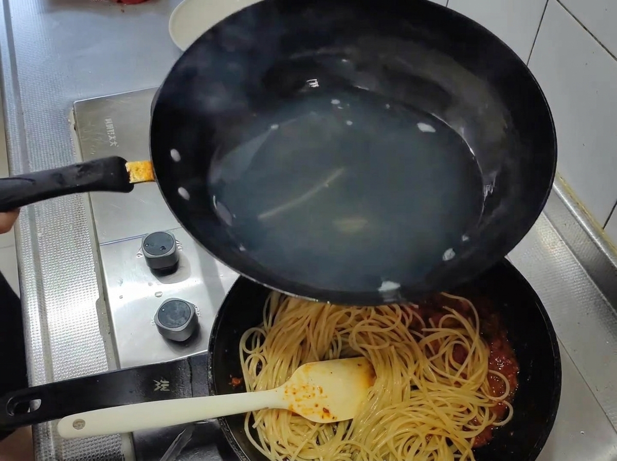 Cooked spaghetti being moved from the boiling water into a pan with a rich red tomato sauce.