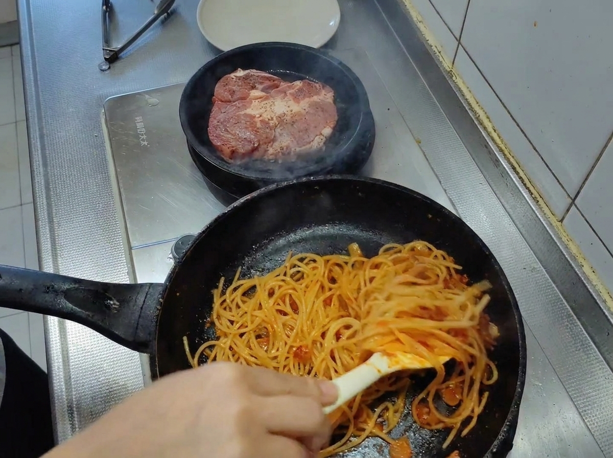 Spaghetti being tossed with tongs in a pan, becoming fully coated in a thick, vibrant tomato sauce.