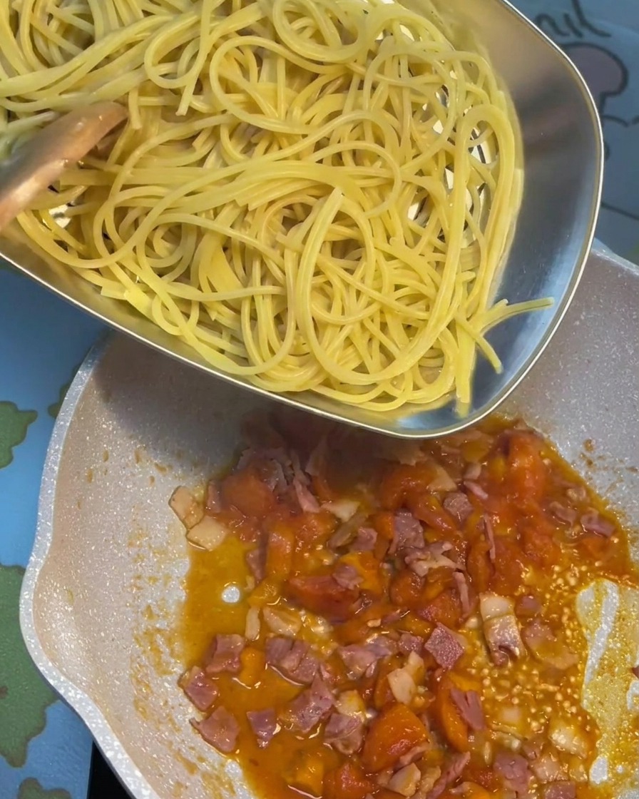 Cooked pasta being tossed in a pan with a rich tomato and bacon sauce.