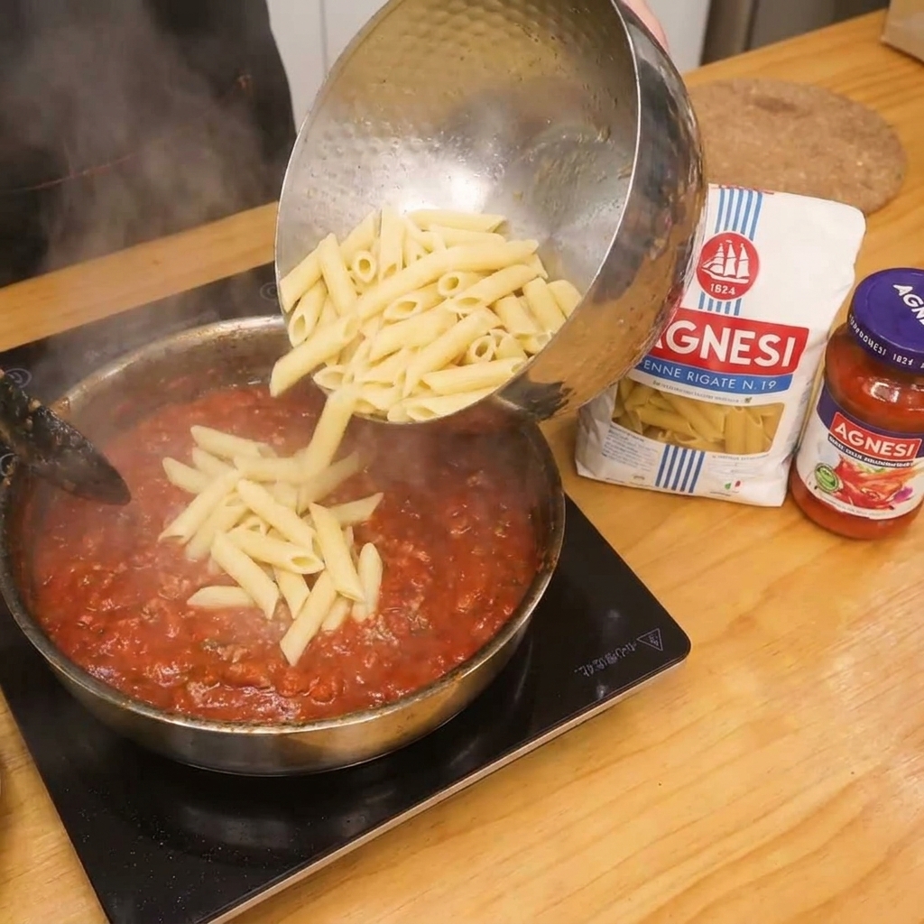 Penne pasta being added to the tomato bacon sauce in the pan for final mixing.