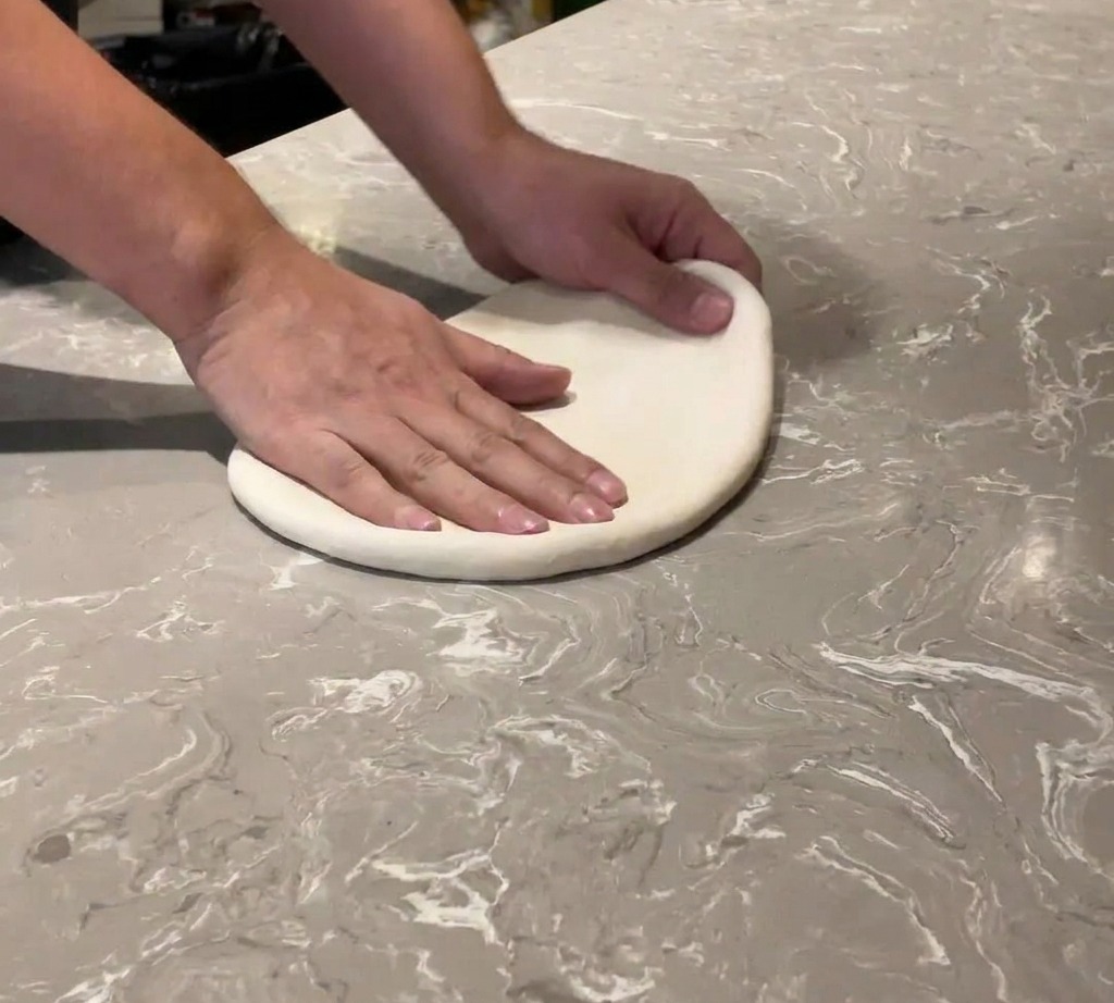 Close-up of hands stretching a soft ball of pizza dough on a marble surface.