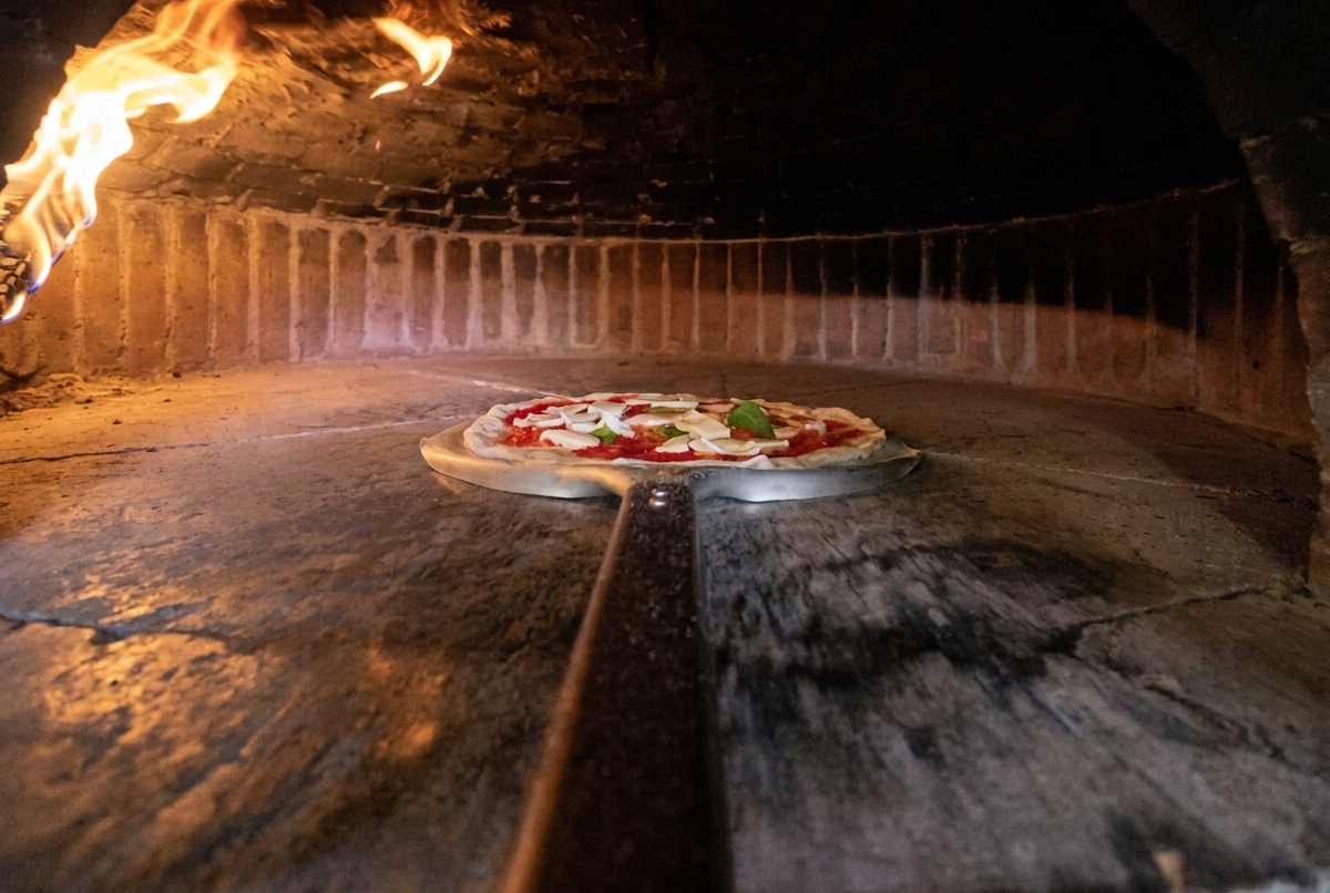 A long metal peel transferring an assembled raw pizza into a hot, glowing wood-fired oven.