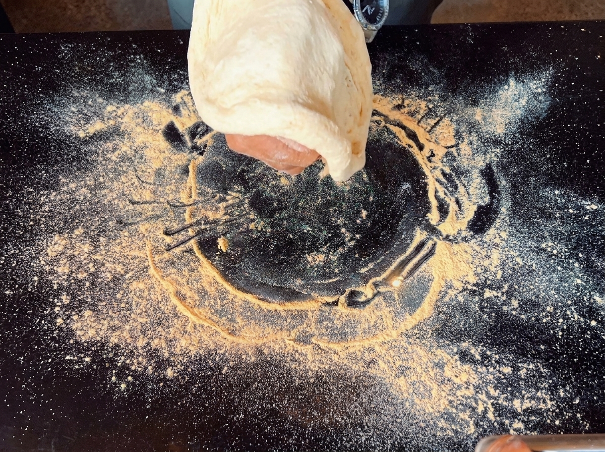 A hand stretching a piece of pizza dough in the air above a flour-dusted black surface.
