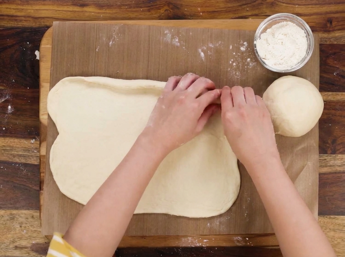 Hands folding the edges of a dough oval to form a boat-like shape, with a bowl of shredded cheese nearby.