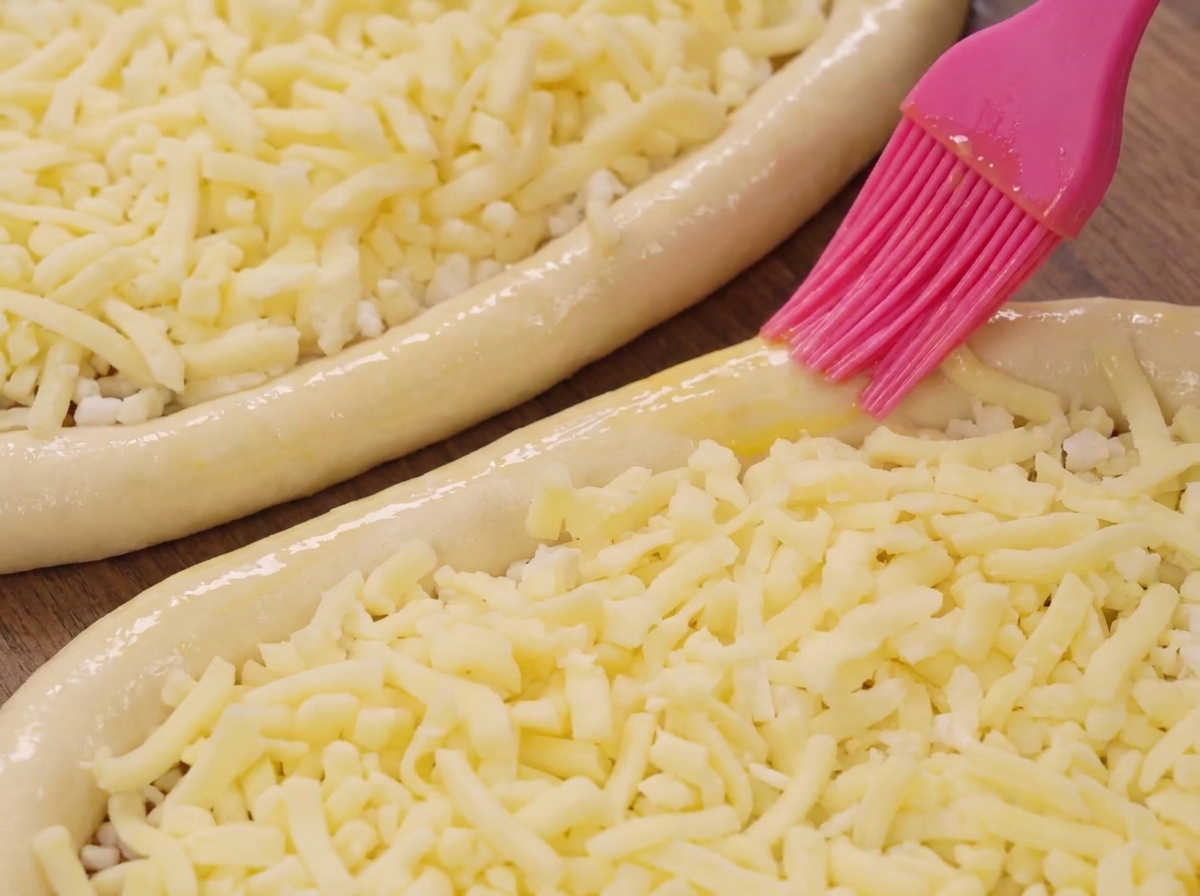 A pink silicone brush applying oil to the edge of a dough boat filled with mozzarella cheese.