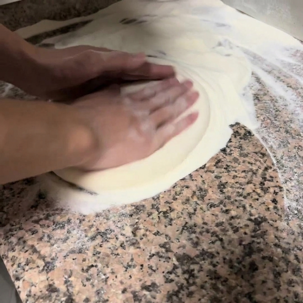 Hands gently pressing and stretching a round pizza dough on a floured countertop.