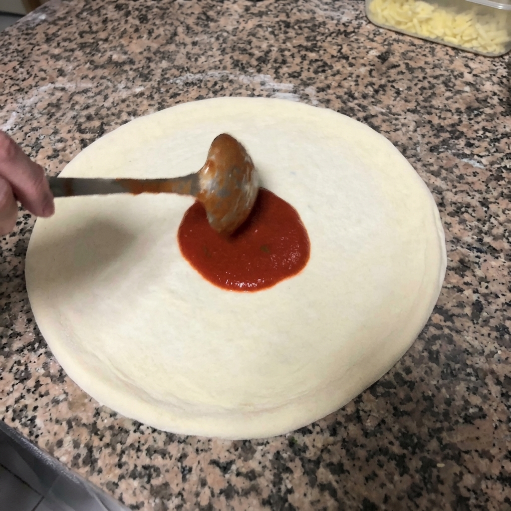 Spreading rich red tomato sauce over the pizza dough in a circular motion using a metal ladle.