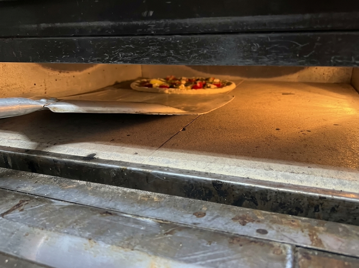 Sliding an assembled fresh pizza off a metal peel into a glowing hot oven onto a baking stone.