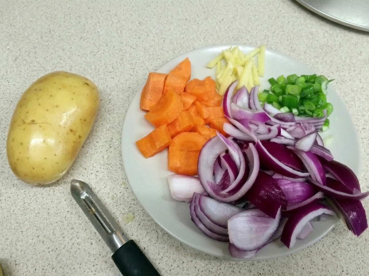 A white plate holding chopped carrots, red onions, ginger strips, and scallions next to a raw potato and a peeler.