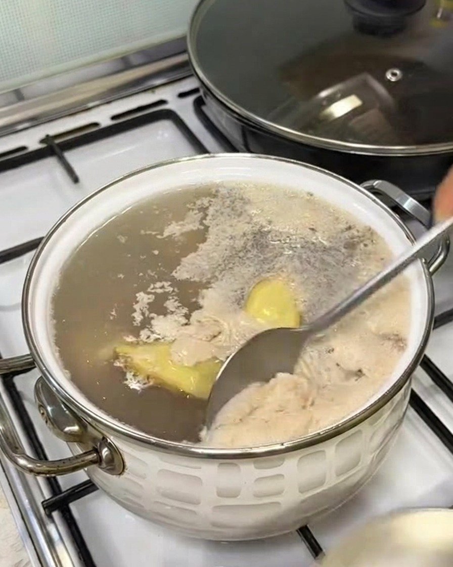 A spoon skimming grayish foam off the surface of boiling water in a pot.