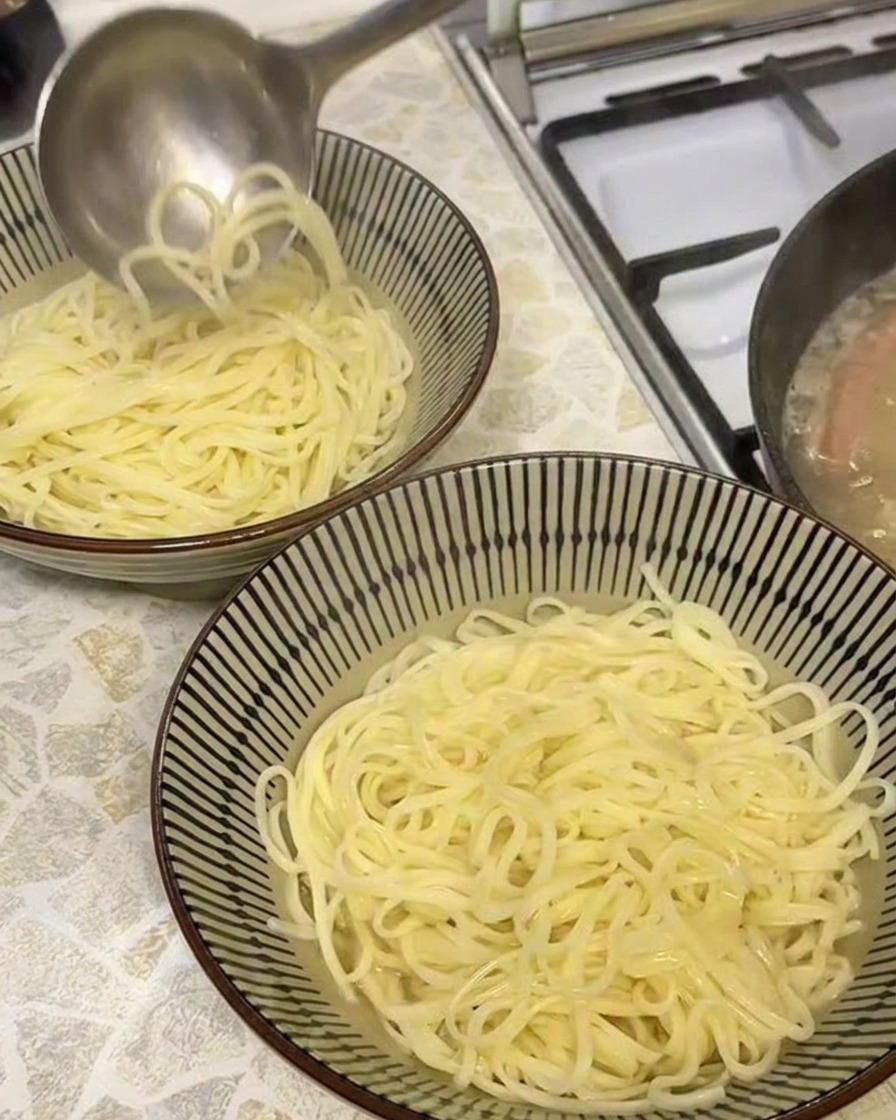 A ladle pouring steaming, opaque golden pork broth into a bowl containing freshly boiled ramen noodles.