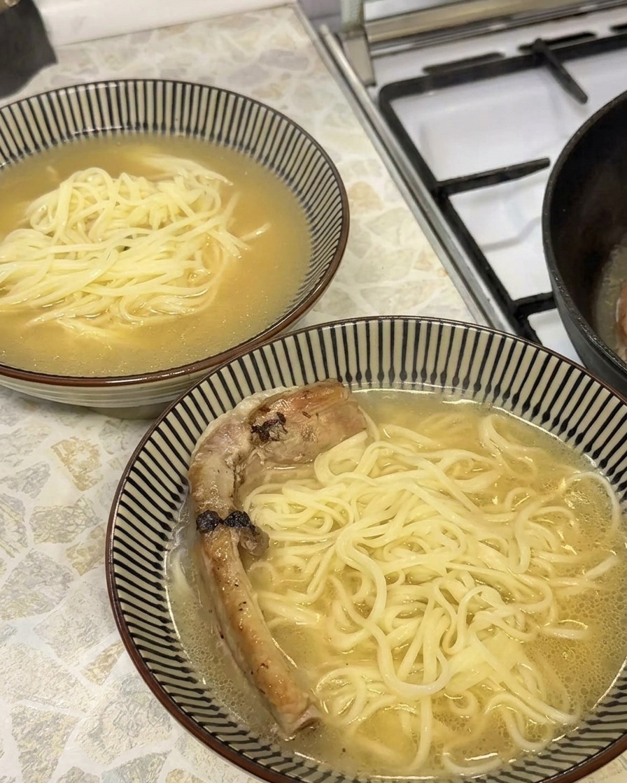 A close-up of a tender, golden-brown braised pork rib being placed into a bowl of ramen broth and noodles.