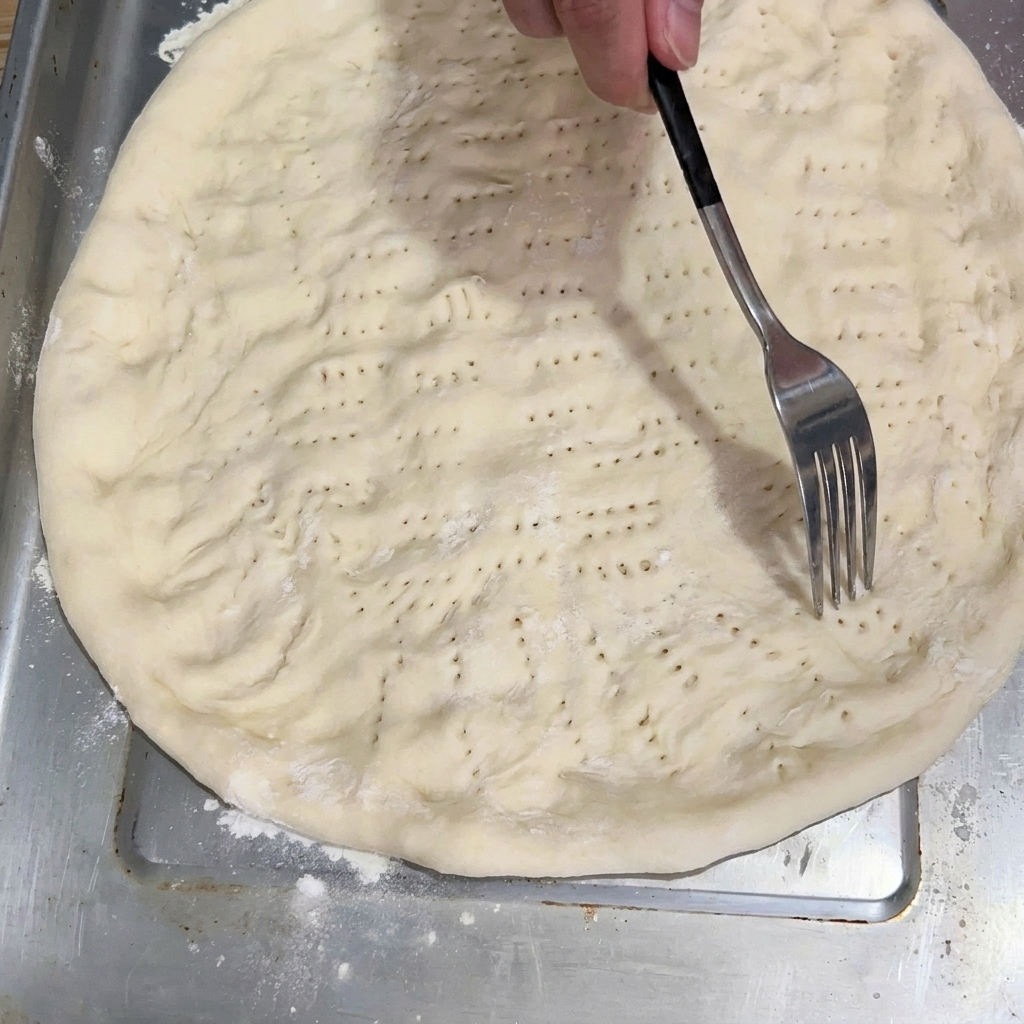 A fork pricking the flattened dough base on the baking tray.