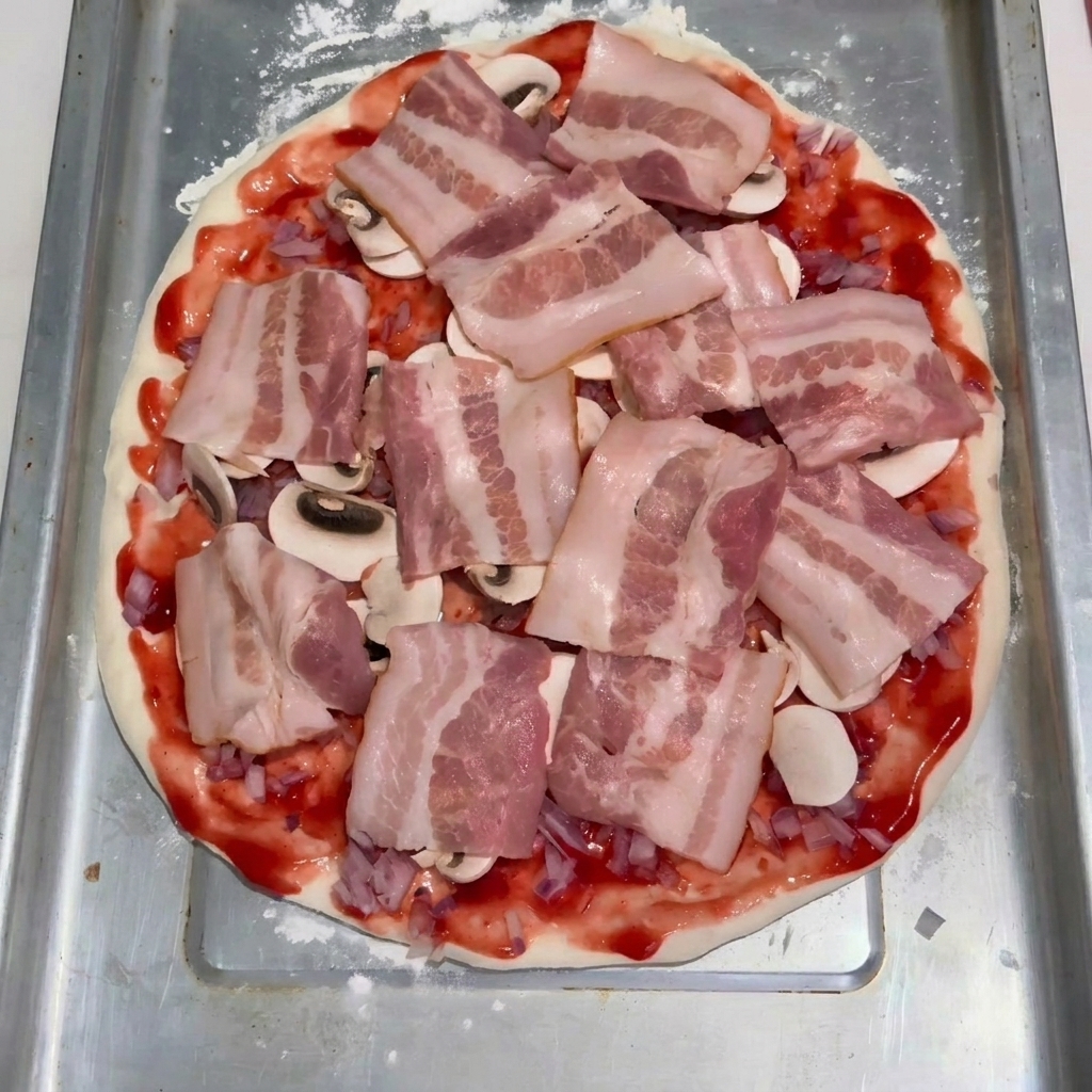 Slices of raw bacon arranged neatly over a layer of sliced mushrooms and tomato sauce on a flattened pizza dough base.
