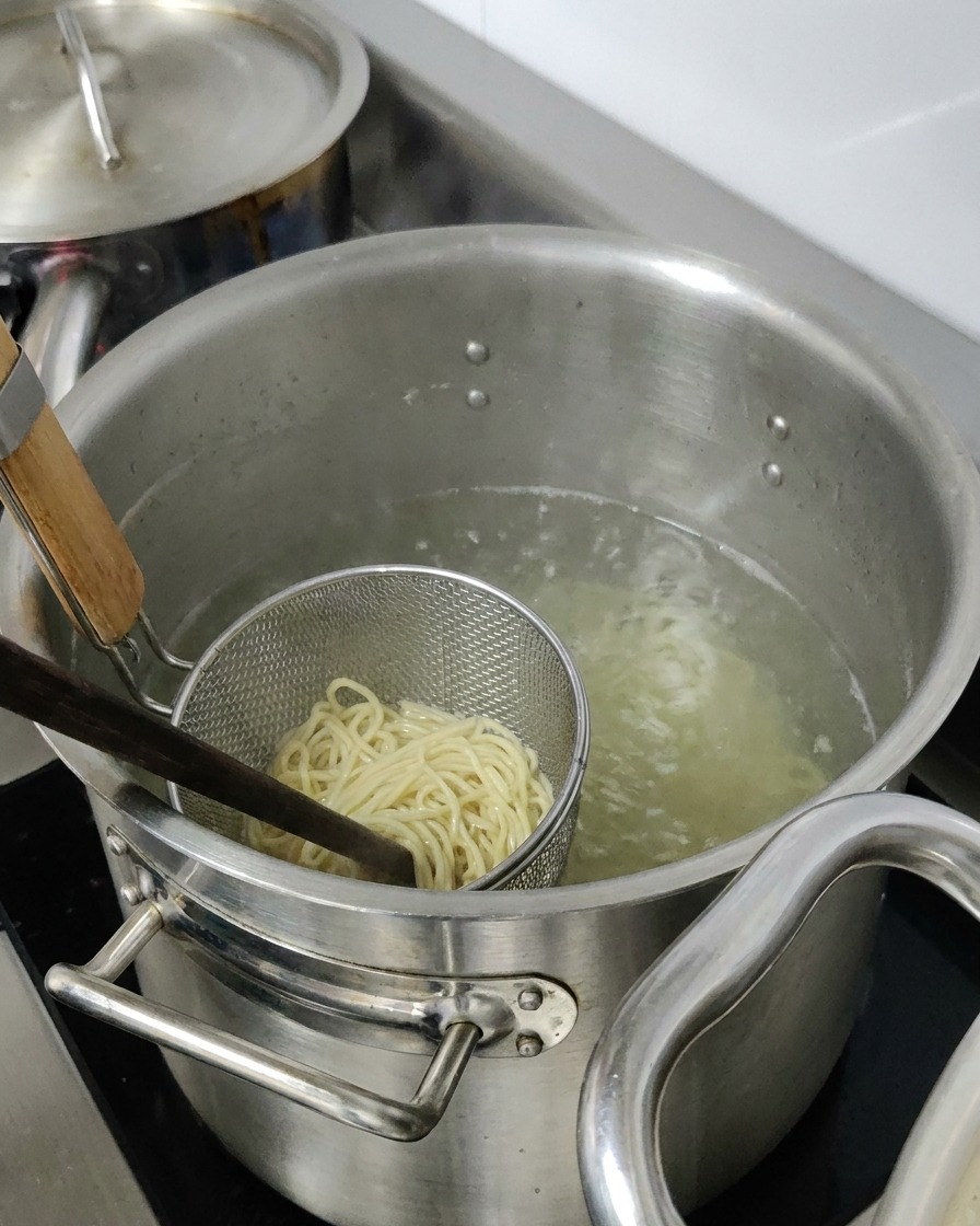 Ramen noodles boiling inside a metal strainer basket submerged in a large pot of hot water.