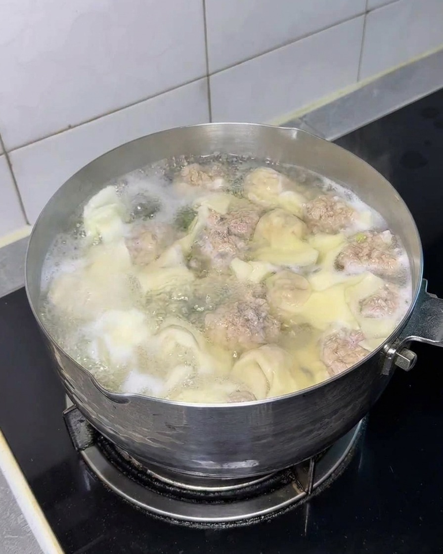 A square block of dry instant noodles submerged in the pot with wontons and meat balls.