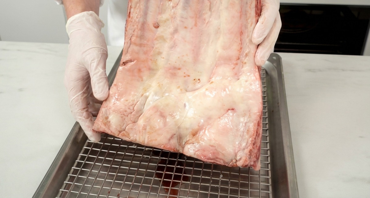 A large piece of raw, red beef ribs with visible white fat marbling held by a chef over a metal rack.