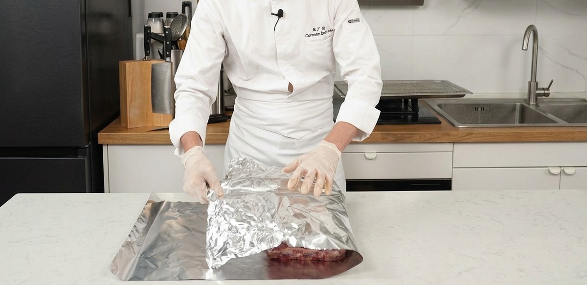 A chef folding a large sheet of silver aluminum foil over seasoned beef ribs on a kitchen counter.