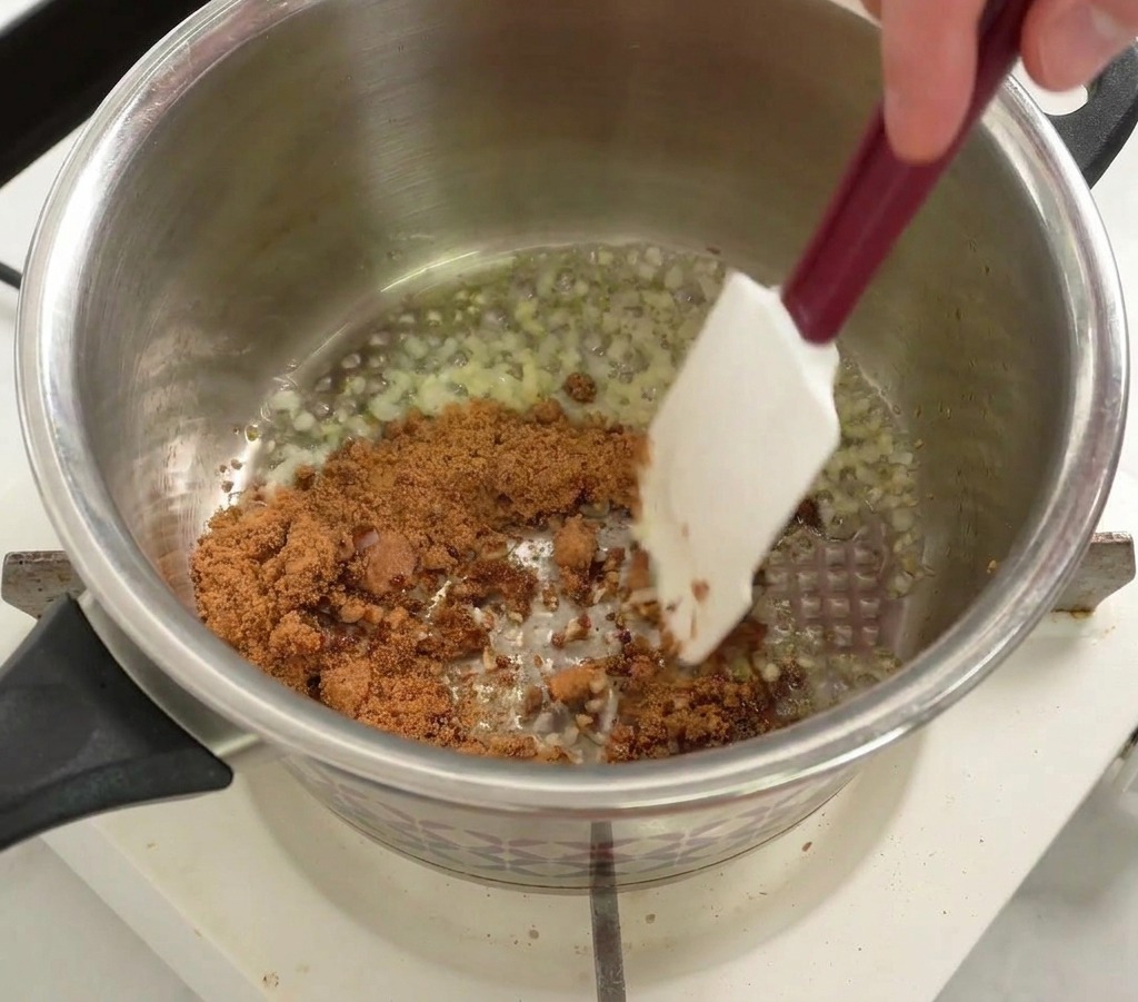 Brown sugar being stirred into a stainless steel pot with sautéed onions and garlic using a white spatula.