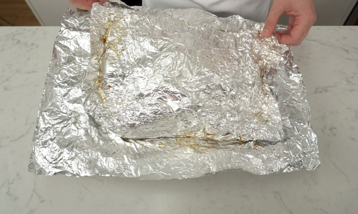 A pair of hands carefully peeling back crinkled aluminum foil to reveal a large slab of slow-roasted beef ribs on a white countertop.