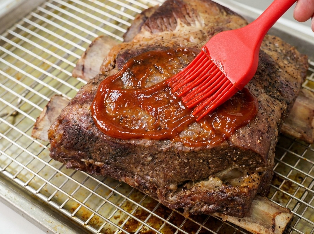 A hand using a red silicone brush to spread a thick, glossy BBQ sauce over a large slab of cooked beef ribs on a wire rack.