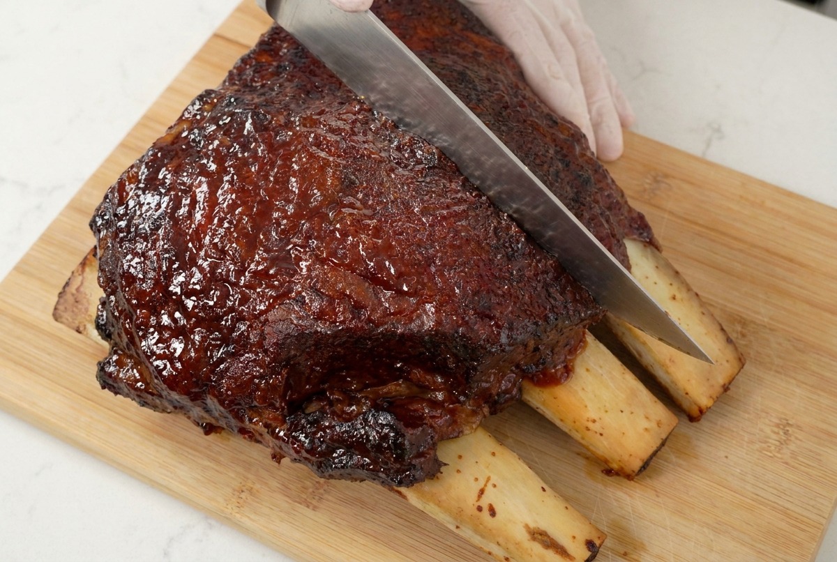 A sharp chefs knife slicing through a dark, caramelized slab of beef ribs on a wooden board, showing the juicy meat texture.