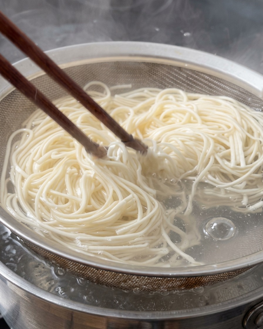 Fresh ramen noodles being lifted from boiling water in a metal wire strainer using wooden chopsticks.