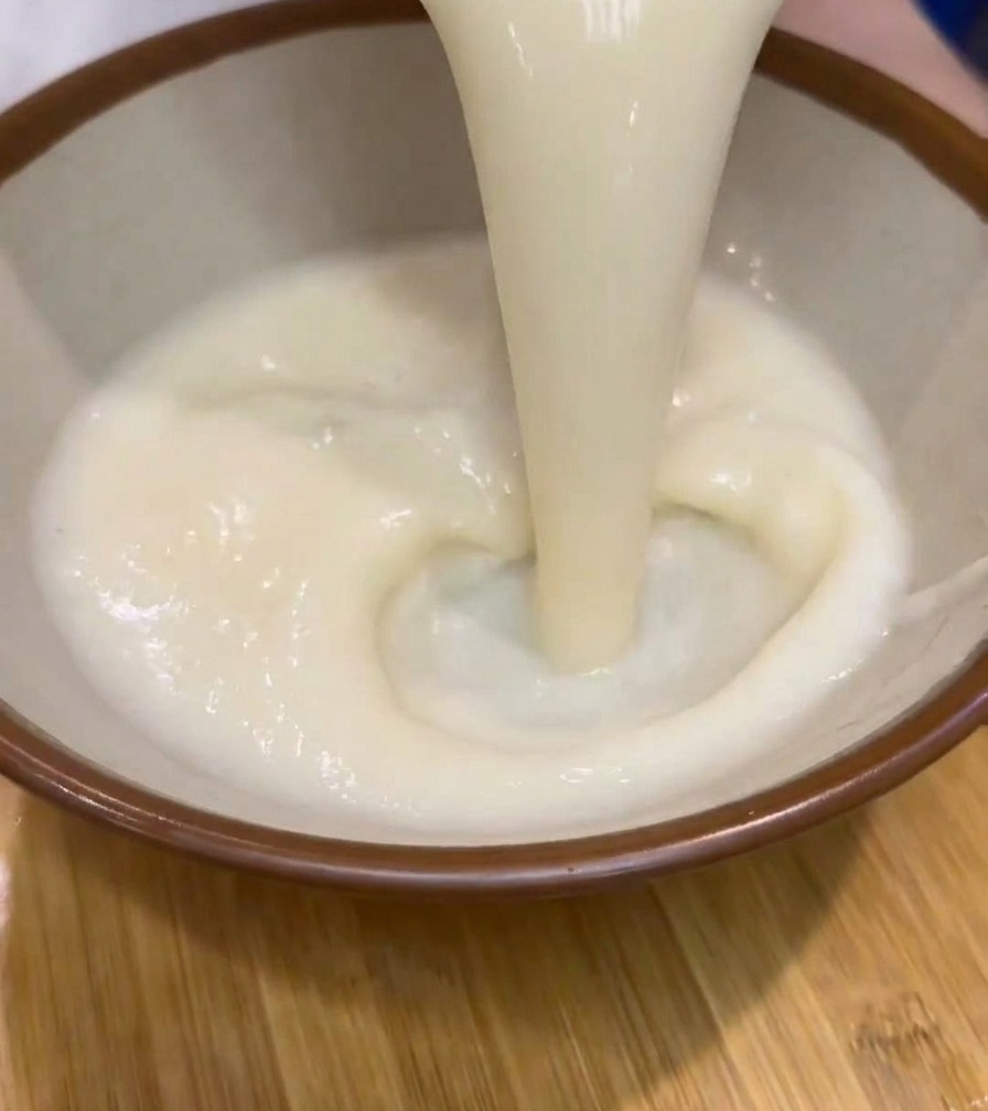 Thick, creamy white hot ramen soup being smoothly poured from a pitcher into a brown-rimmed ceramic serving bowl.