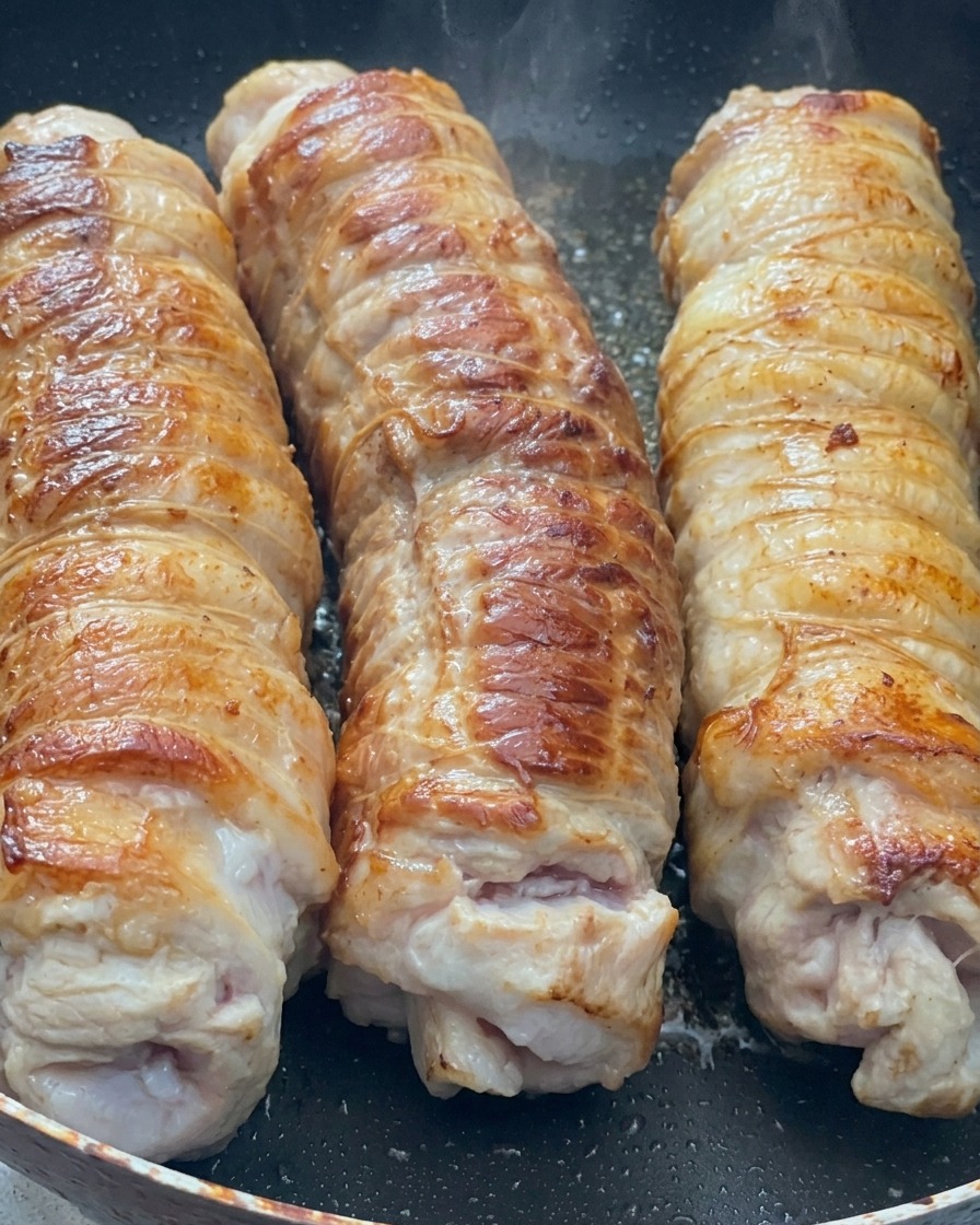 Three large rolls of tied pork belly searing in a hot pan, showing a golden-brown, caramelized crust.