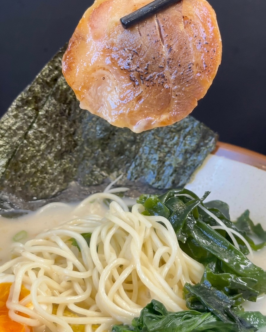 Black chopsticks lifting a caramelized slice of char siu pork over a fully assembled bowl of ramen filled with noodles, broth, seaweed, and a soft-boiled egg.