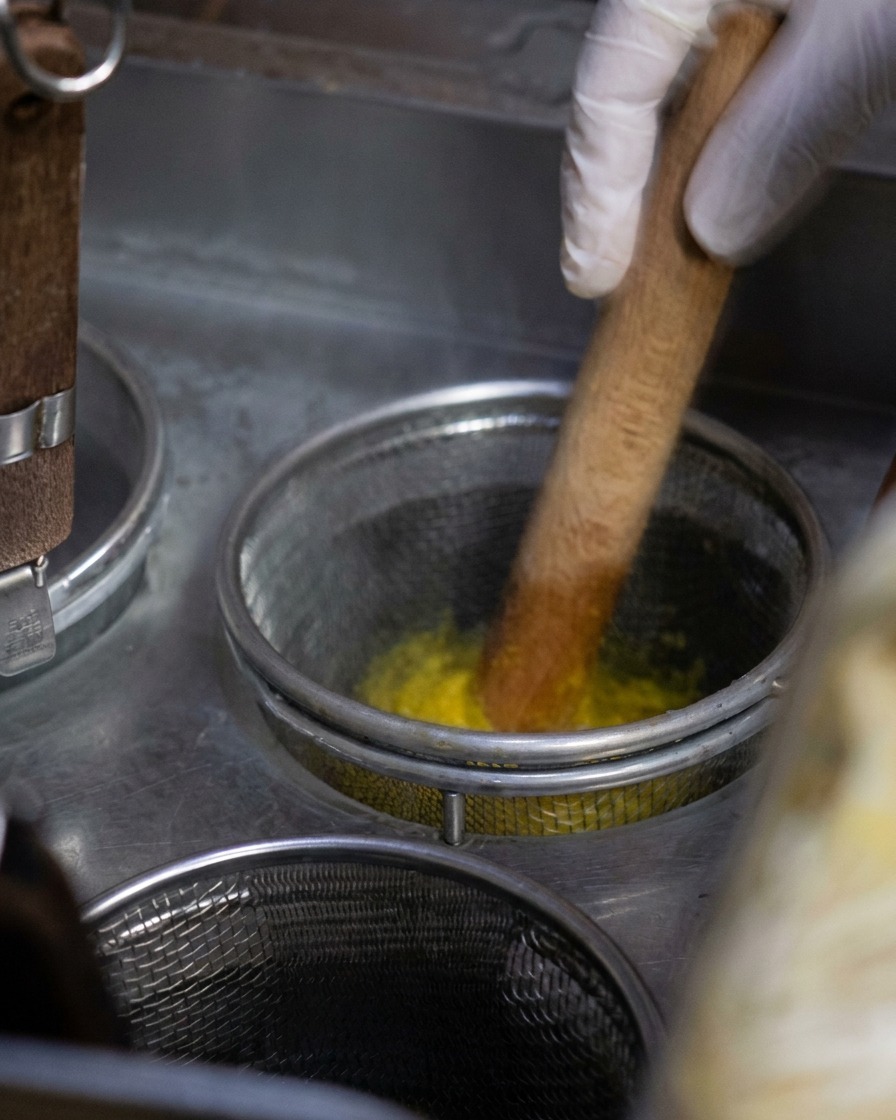 A metal mesh basket filled with yellow noodles being stirred with a wooden tool in a pot of vigorously boiling water.