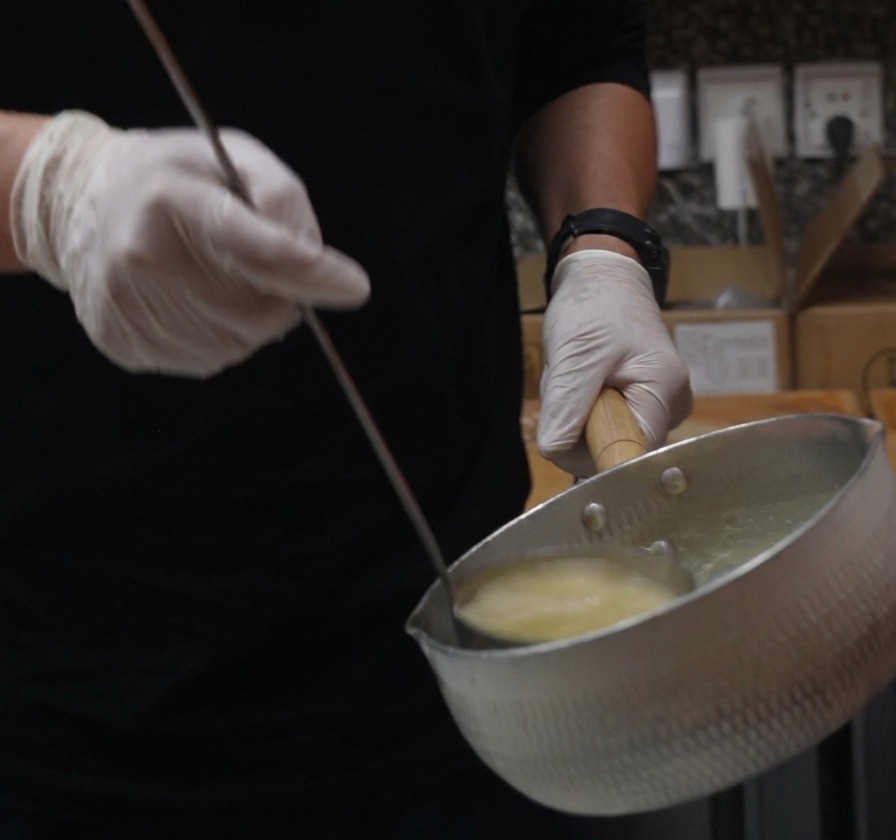 Gloved hands holding a metal saucepan and a ladle, pouring creamy yellow chicken paitan broth.