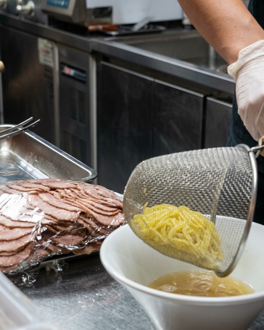 A metal strainer dropping freshly cooked yellow ramen noodles into a white bowl filled with creamy broth.
