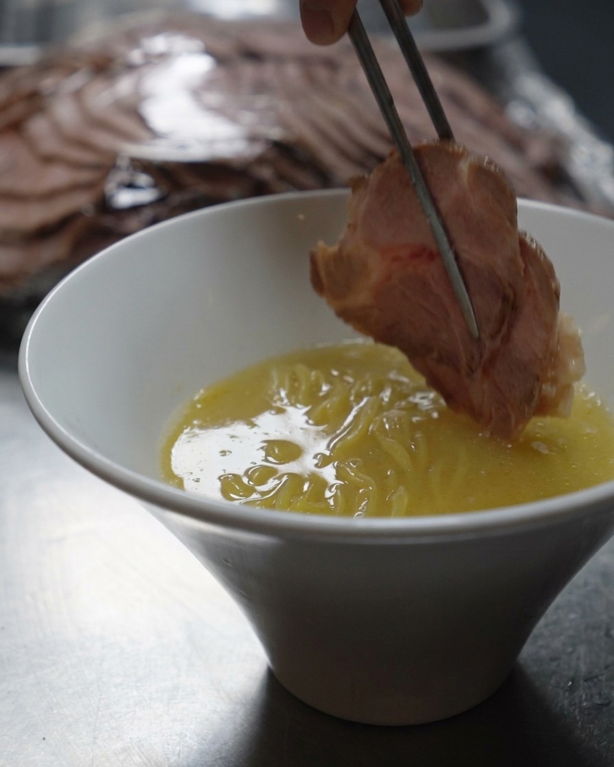Metal chopsticks lowering a thick slice of seared pork into a bowl of ramen noodles and creamy broth.