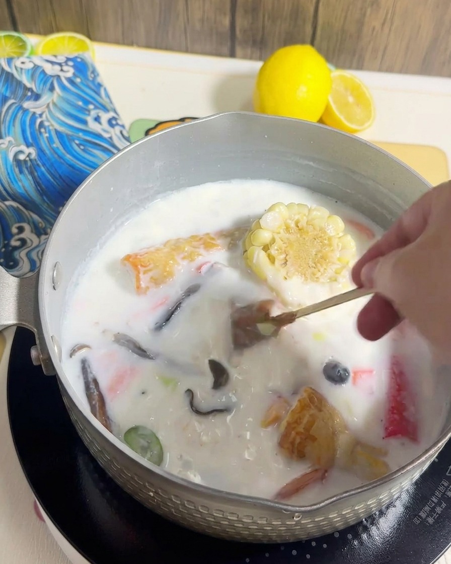 Ramen noodles being added to a pot filled with creamy broth and various cooked toppings.