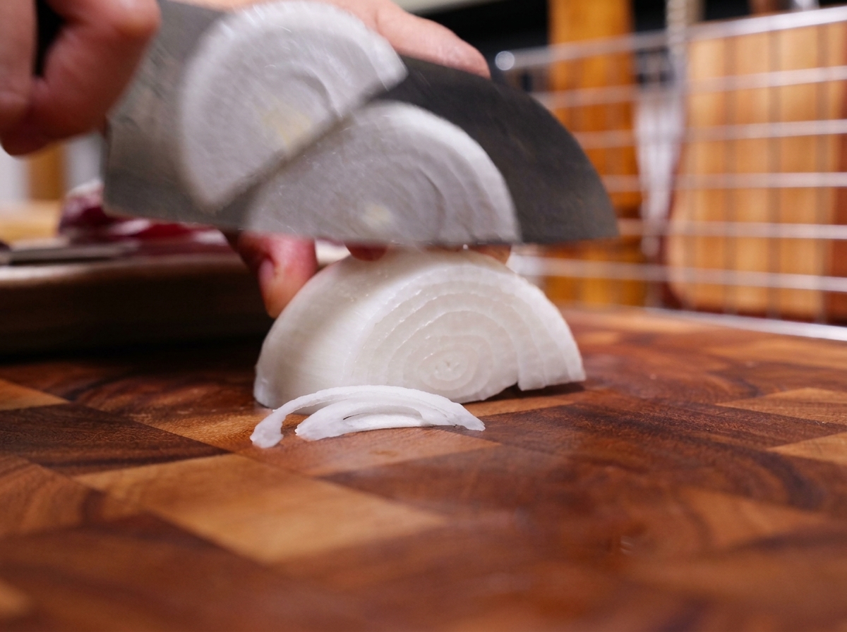 A chefs knife slicing a white onion half into thin strips on a wooden cutting board.