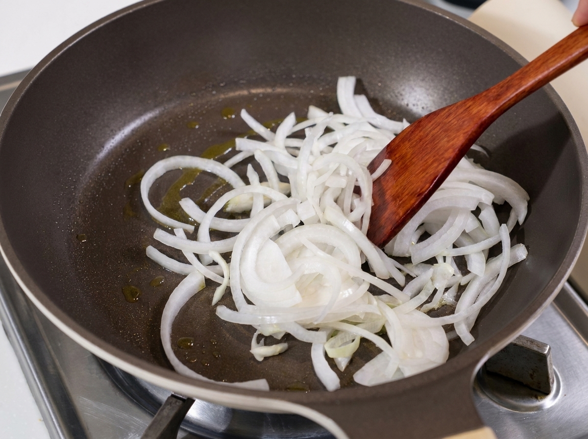 Sliced white onions being sautéed in a pan with oil using a wooden spatula.