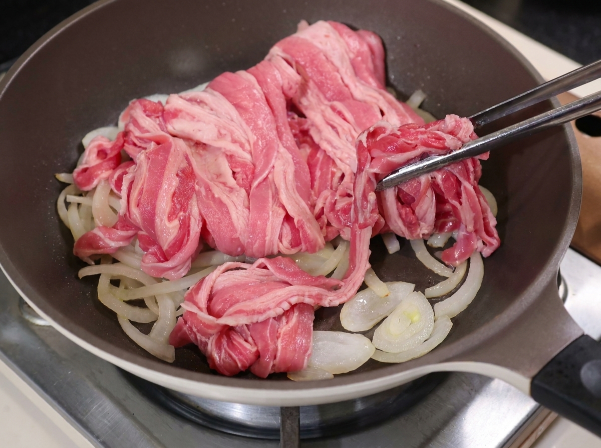 Metal tongs placing raw, thinly sliced fatty beef on top of cooked onions in a hot pan.